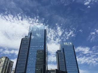 Tall skyscrapers with reflective glass windows rise against a backdrop of a clear blue sky with scattered clouds. The buildings feature modern architecture, with the sign 'Bruno Central Plaza' visible on one of them.