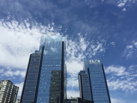 Tall skyscrapers with reflective glass windows rise against a backdrop of a clear blue sky with scattered clouds. The buildings feature modern architecture, with the sign 'Bruno Central Plaza' visible on one of them.