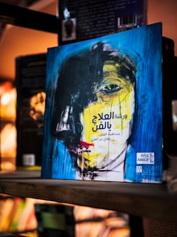 A book with a striking abstract cover featuring a distorted face painted with bold brush strokes in blue, black, and yellow. The background blurs into a shelf of books, creating a warm and cozy atmosphere.