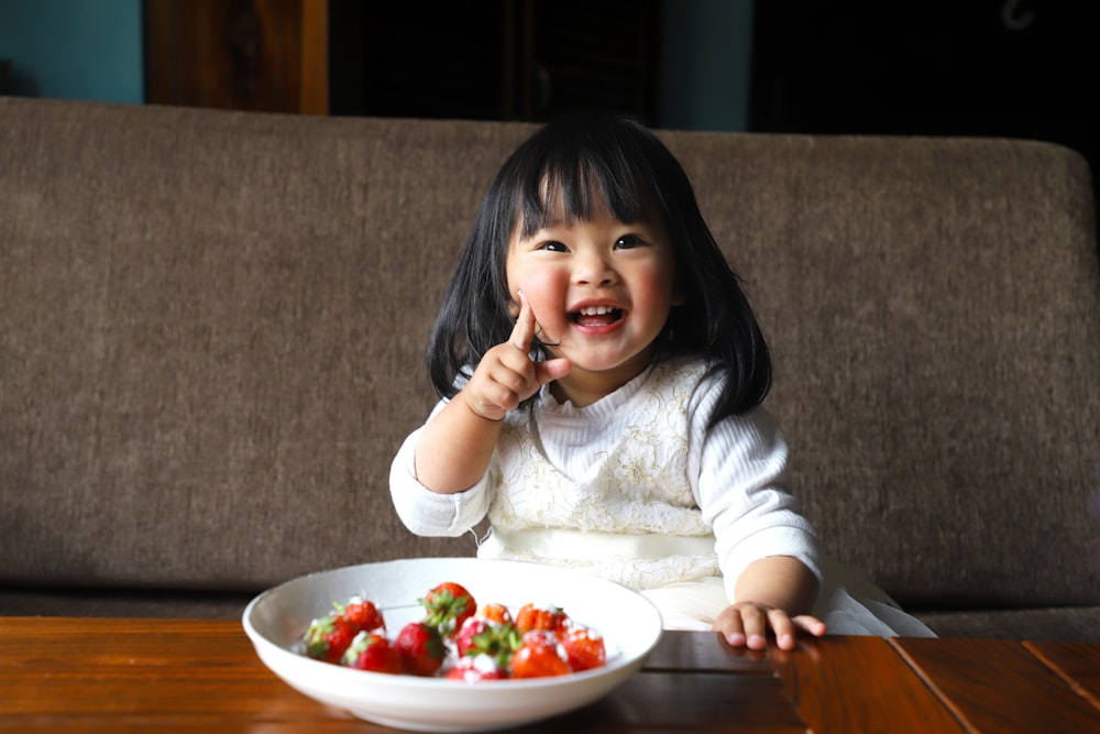 a little girl sitting in front of a bowl of fruit