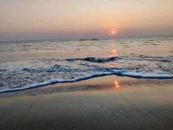 The image depicts a serene beach scene during sunset with gentle waves lapping onto the shore. The sun is low on the horizon, casting a warm glow across the water and sky. The smooth sand reflects the colors of the sunset.