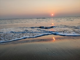 The image depicts a serene beach scene during sunset with gentle waves lapping onto the shore. The sun is low on the horizon, casting a warm glow across the water and sky. The smooth sand reflects the colors of the sunset.