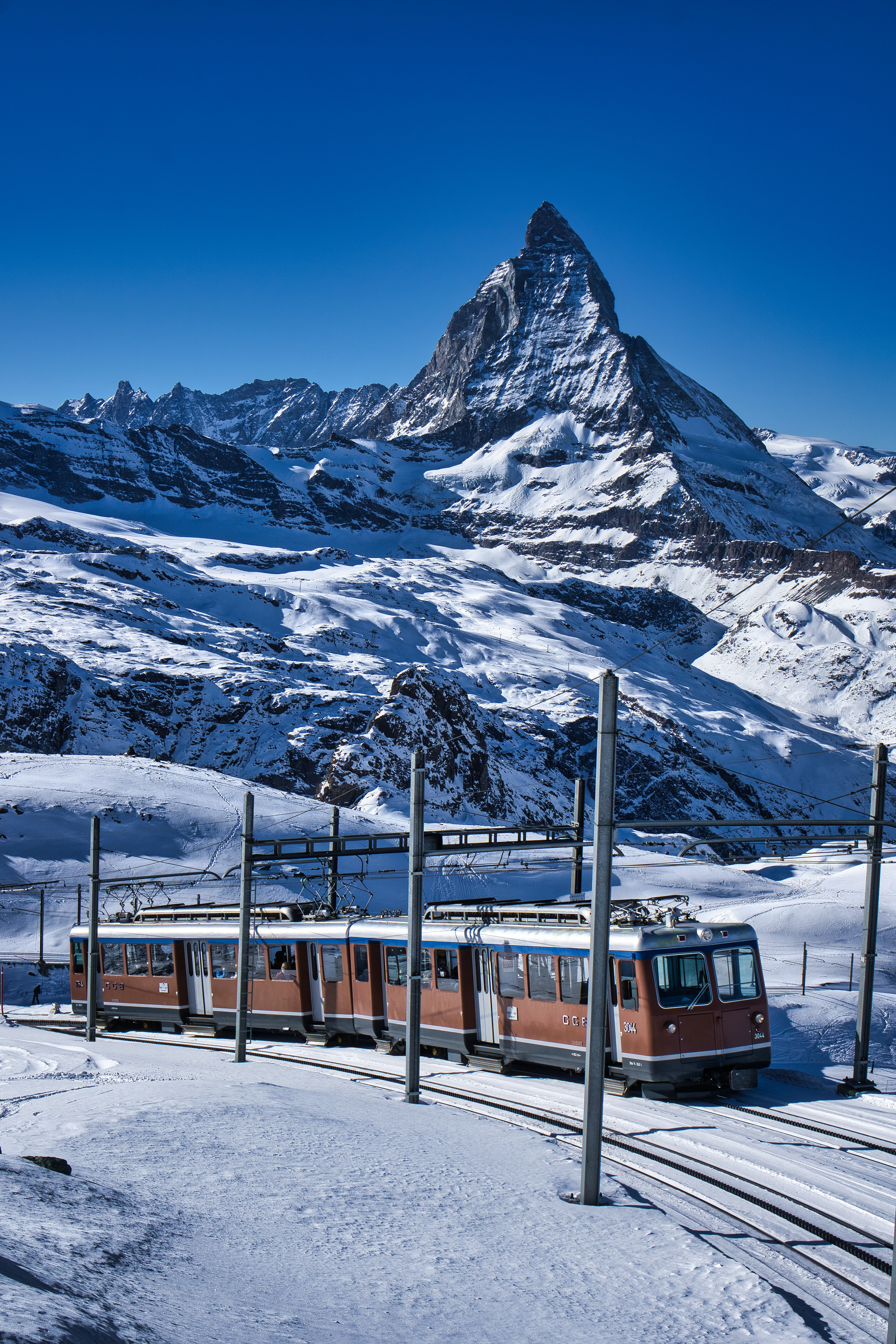 A red Gornergrat cog railway train standing on snow-covered track at the summit station above Zermatt, the dark pyramid of the Matterhorn rising into a cloudless deep-blue sky behind it.
