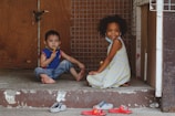 Two young children are sitting on a concrete doorstep in front of a wooden and wire mesh door. One child is wearing a blue and red shirt with jeans and is eating a snack. The other child, dressed in a light-colored patterned dress, is smiling at the camera. Both children are barefoot, with a few pairs of colorful sandals and slippers scattered on the ground in front of them.