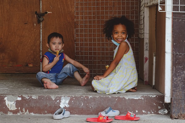 Two young children are sitting on a concrete doorstep in front of a wooden and wire mesh door. One child is wearing a blue and red shirt with jeans and is eating a snack. The other child, dressed in a light-colored patterned dress, is smiling at the camera. Both children are barefoot, with a few pairs of colorful sandals and slippers scattered on the ground in front of them.