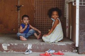 Two young children are sitting on a concrete doorstep in front of a wooden and wire mesh door. One child is wearing a blue and red shirt with jeans and is eating a snack. The other child, dressed in a light-colored patterned dress, is smiling at the camera. Both children are barefoot, with a few pairs of colorful sandals and slippers scattered on the ground in front of them.