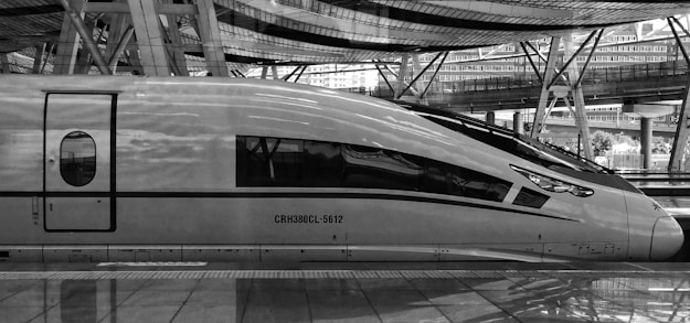 A sleek high-speed train is stationed on the platform, featuring an aerodynamic design with large windows and streamlined contours. The background comprises a modern railway station with a complex structure of glass and steel, emphasizing an urban and futuristic setting.