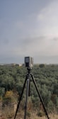 A camera or surveying device is mounted on a tripod, facing a vast expanse of olive groves that stretch towards the horizon. The sky is overcast, contributing to a subdued atmosphere. A few scattered buildings are visible in the distance.