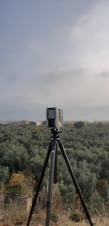 A camera or surveying device is mounted on a tripod, facing a vast expanse of olive groves that stretch towards the horizon. The sky is overcast, contributing to a subdued atmosphere. A few scattered buildings are visible in the distance.
