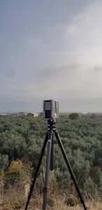 A camera or surveying device is mounted on a tripod, facing a vast expanse of olive groves that stretch towards the horizon. The sky is overcast, contributing to a subdued atmosphere. A few scattered buildings are visible in the distance.