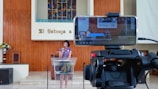 A woman is speaking at a podium inside a church, with a smartphone on a tripod in the foreground recording the event. The background features a colorful stained glass window and wooden paneling with a Spanish phrase. There are flowers on the left side and audio equipment visible.