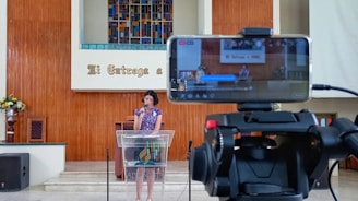 A woman is speaking at a podium inside a church, with a smartphone on a tripod in the foreground recording the event. The background features a colorful stained glass window and wooden paneling with a Spanish phrase. There are flowers on the left side and audio equipment visible.