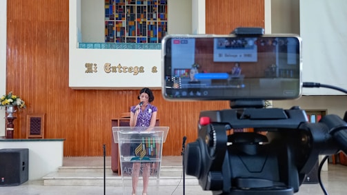 A woman is speaking at a podium inside a church, with a smartphone on a tripod in the foreground recording the event. The background features a colorful stained glass window and wooden paneling with a Spanish phrase. There are flowers on the left side and audio equipment visible.