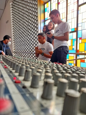 A group of people are gathered around a large audio mixing console. One person is wearing a mask and appears to be operating the equipment. The others are observing or interacting with the console. They are standing in front of a colorful stained glass window that casts vibrant light into the room.