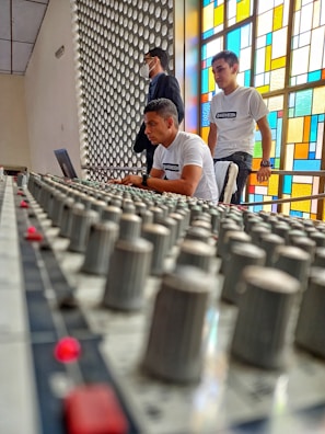 A close-up view of a sound mixing console with multiple knobs in the foreground. Behind the console, three people are visible, with two wearing white shirts standing and observing while one is seated and working on a laptop. The scene is set in a bright room with a colorful stained glass window on the right and a decorative wall panel on the left.
