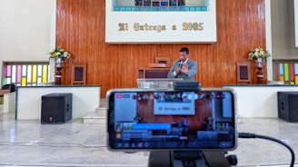 A man is standing on a stage inside a religious venue, possibly a church, delivering a speech or sermon. A smartphone on a tripod in the foreground is recording or streaming the event. The background features a wooden panel with the text 'Mi Entrega a DIOS', along with flower arrangements on both sides and colored glass windows.