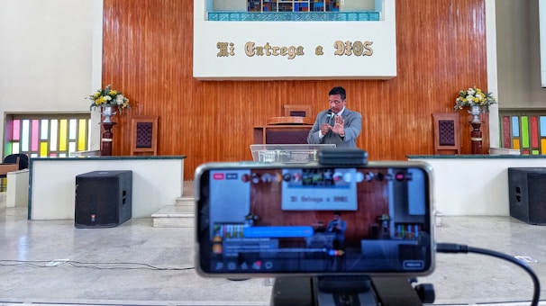 A man is standing on a stage inside a religious venue, possibly a church, delivering a speech or sermon. A smartphone on a tripod in the foreground is recording or streaming the event. The background features a wooden panel with the text 'Mi Entrega a DIOS', along with flower arrangements on both sides and colored glass windows.