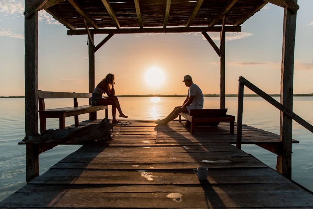 a man and a woman sitting on a dock at sunset
