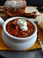 A bowl of hearty chili topped with a dollop of sour cream and a garnish of fresh chives. The chili is rich in color with visible chunks of meat and vegetables. There is a loaf of crusty bread and several slices of it placed on a wooden board in the background.