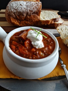 A bowl of hearty chili topped with a dollop of sour cream and a garnish of fresh chives. The chili is rich in color with visible chunks of meat and vegetables. There is a loaf of crusty bread and several slices of it placed on a wooden board in the background.