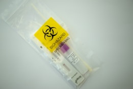A biohazard specimen bag contains a swab and a test cassette for rapid testing. The bag is transparent with a vivid yellow label featuring a biohazard symbol.