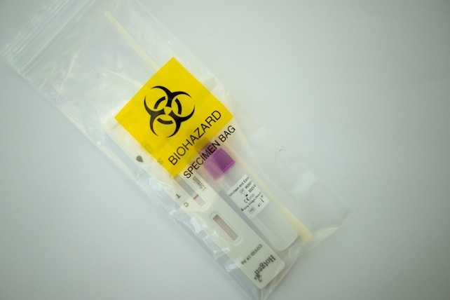 A biohazard specimen bag contains a swab and a test cassette for rapid testing. The bag is transparent with a vivid yellow label featuring a biohazard symbol.