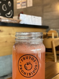 A glass jar filled with a frothy pink beverage labeled 'Frutos Prohibidos' is placed on a wooden table in a cozy café environment. The background features wooden furniture, a chalkboard with fruit illustrations, and a counter with stacked items. The lighting is warm and inviting.