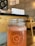 A glass jar filled with a frothy pink beverage labeled 'Frutos Prohibidos' is placed on a wooden table in a cozy café environment. The background features wooden furniture, a chalkboard with fruit illustrations, and a counter with stacked items. The lighting is warm and inviting.