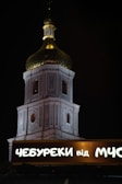 A brightly illuminated church tower with intricate architectural details and a golden dome is visible against a night sky. In the foreground, a lit sign with Cyrillic script reads 'ЧЕБУРЕКИ від МУС'.