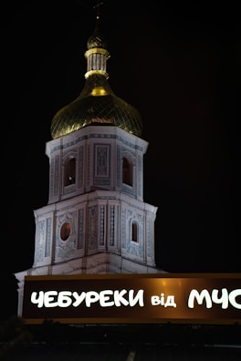 A brightly illuminated church tower with intricate architectural details and a golden dome is visible against a night sky. In the foreground, a lit sign with Cyrillic script reads 'ЧЕБУРЕКИ від МУС'.