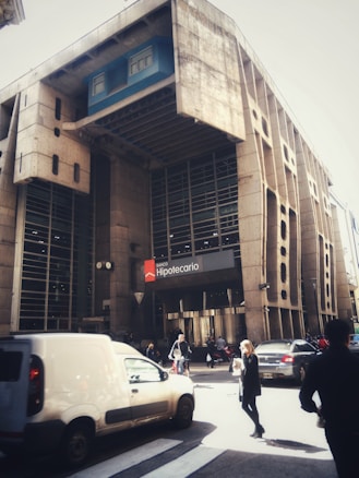 A large, concrete building with an imposing architectural style features a prominent sign for Banco Hipotecario. The frontage displays vertical columns and large glass windows. Pedestrians and vehicles, including a white van and several cars, are visible on the street.