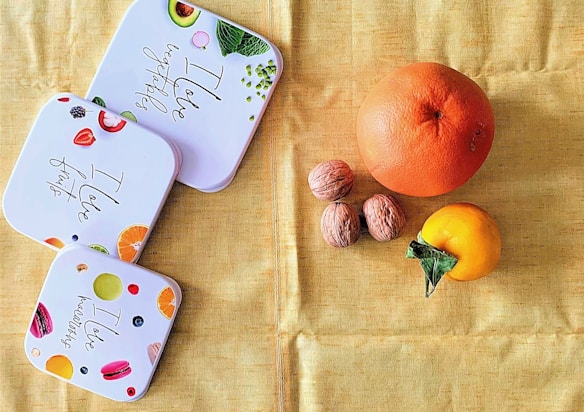 A neatly arranged setup displays three white square containers decorated with images of fruits, vegetables, and macarons. Each container features text such as 'I love fruits' and 'I love vegetables'. To the right, there is a large grapefruit, three walnuts, and a persimmon resting on a soft, golden-yellow fabric background.