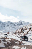 A convoy of 4x4 vehicles crossing a rocky mountain pass in Ladakh with snow-capped peaks in the background.