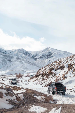 A convoy of 4x4 vehicles crossing a rocky mountain pass in Ladakh with snow-capped peaks in the background.