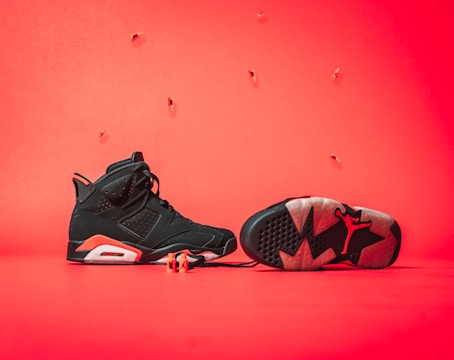 A pair of black and red athletic shoes is displayed against a bright red background. One shoe is upright showing the side profile, while the other is lying on its side revealing the sole with a distinctive logo. The laces are partially visible, and small decorative elements are suspended in the air.