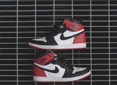 A pair of sneakers with a black, white, and red color scheme is arranged horizontally against a metal grate. The sneakers feature a recognizable logo and appear to be of a high-top design. The metal grate forms a grid-like pattern in the background.