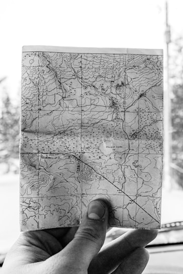 A black and white photograph of a close-up hand holding a detailed topographical map. The map displays contour lines, bodies of water, and textual labels of locations such as Thayer Lake. The background is blurred, suggesting the photo was taken inside a vehicle.