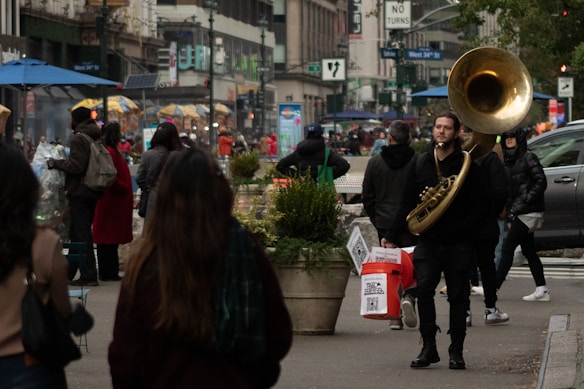 A busy urban street scene with people walking and a musician playing a large brass instrument. There are several food stalls with umbrellas in the background, and buildings line the street.