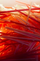 A close-up, chaotic arrangement of plastic drinking straws in various shades of red, orange, and yellow. The straws are layered and intertwined, creating a visually striking pattern with a warm, glowing light effect.