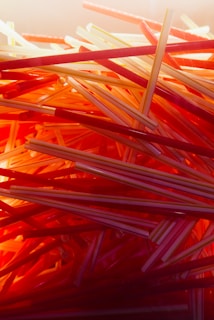 A close-up, chaotic arrangement of plastic drinking straws in various shades of red, orange, and yellow. The straws are layered and intertwined, creating a visually striking pattern with a warm, glowing light effect.