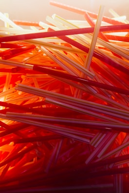 A close-up, chaotic arrangement of plastic drinking straws in various shades of red, orange, and yellow. The straws are layered and intertwined, creating a visually striking pattern with a warm, glowing light effect.