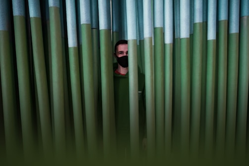 A person wearing a face mask is partially obscured by large green vertical pipes. The scene is dimly lit, creating a sense of secrecy or concealment.
