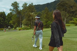 A woman with long hair walks alongside a man carrying golf clubs on a lush green golf course. Trees and hills are in the background, creating a serene, natural setting.
