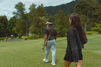 A woman with long hair walks alongside a man carrying golf clubs on a lush green golf course. Trees and hills are in the background, creating a serene, natural setting.
