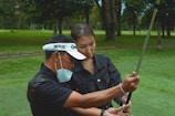 A person wearing a mask and a visor is helping another person with their golf swing on a grassy field surrounded by trees.