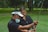 A person wearing a mask and a visor is helping another person with their golf swing on a grassy field surrounded by trees.