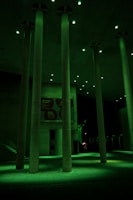 Tall concrete columns are illuminated by green lighting in a modern architectural setting. The space appears to be a public area, possibly an atrium or open hallway, with a high ceiling and visible signs on the walls in the background.