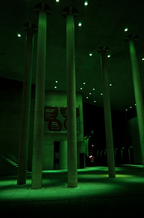 Tall concrete columns are illuminated by green lighting in a modern architectural setting. The space appears to be a public area, possibly an atrium or open hallway, with a high ceiling and visible signs on the walls in the background.