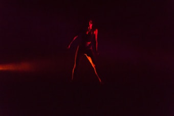 A silhouette of a person in a dynamic pose is surrounded by deep, dark shadows. The lighting casts a dramatic red and orange glow, creating a sense of movement and intensity.
