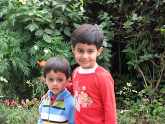 Two young children are standing together in front of a lush green background with various plants and flowers. The older child is wearing a red shirt and has their arm around the younger one, who is wearing a colorful striped sweater.